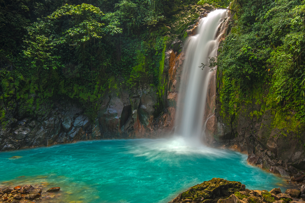 A waterfall with water falling down it into the blue pool.