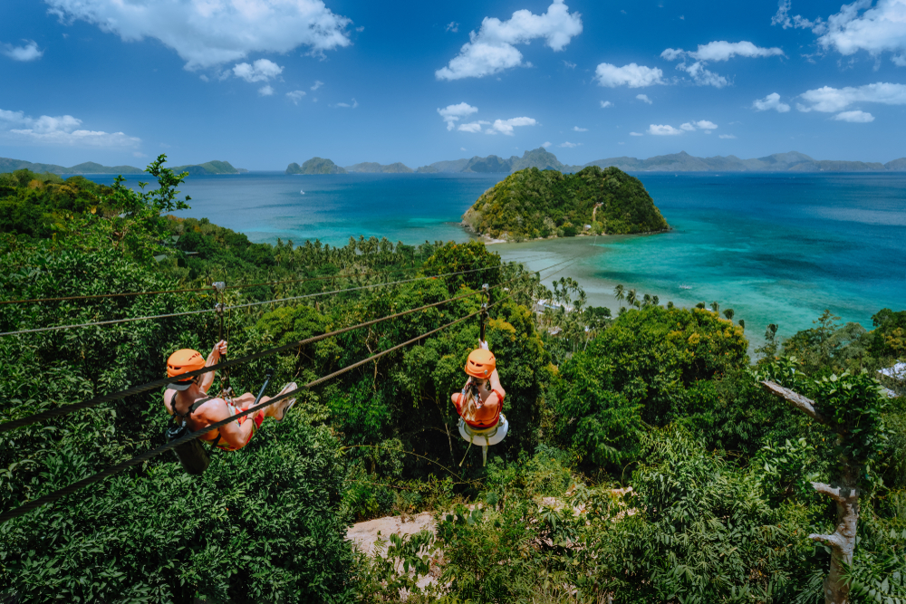 Two people are ziplining over a lush green hillside.